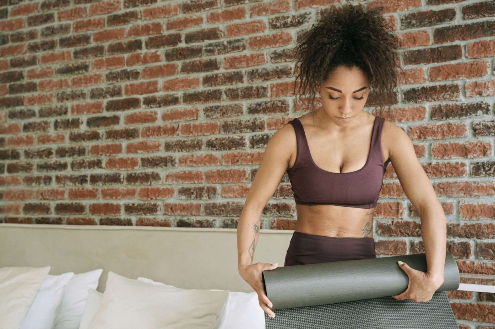 Woman in maroon activewear rolling out a yoga mat in front of an exposed brick wall