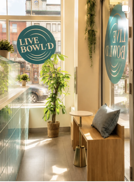 Sunlit Live Bowl'd storefront entryway with teal logo decals on the windows, a leafy plant, and a wooden bench with a grey cushion