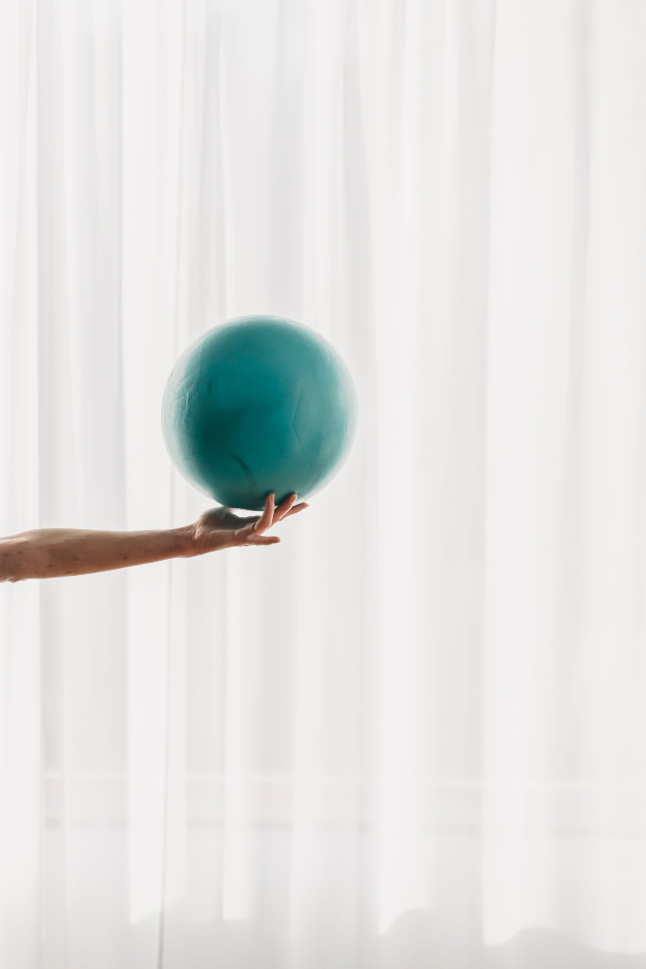 A hand holding a teal pilates ball in front of soft white sheer curtains